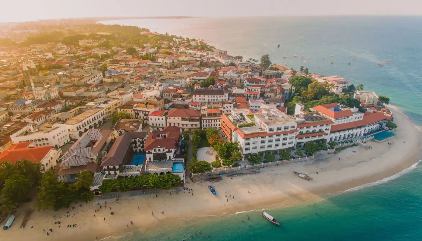 Vue aérienne de Stone Town, toits de la vieille ville, façades blanches face à l'océan et plage avec bateaux au premier plan